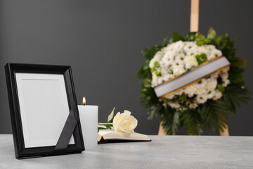 Photo frame with black ribbon, rose, burning candle on light table and wreath of flowers near grey...