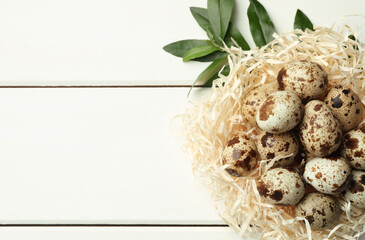 Nest with many speckled quail eggs and green leaves on white wooden table. Space for text