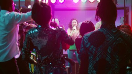 Smiling girl doing karaoke at club, singing songs at microphone and dancing with DJ on stage. Young man mixing music for crowd of people having fun and partying, dance party. Handheld shot.