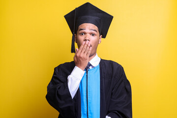 Young man in graduation cap and ceremony robe afraid and shocked, surprise and amazed expression with hands on mouth isolated over yellow background. Omg and wow. 