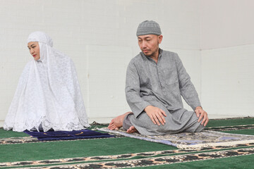 Asian Muslim man becoming imam and leading his wife to pray, gesturing last movement of salat, reciting salam to the right then to the left.