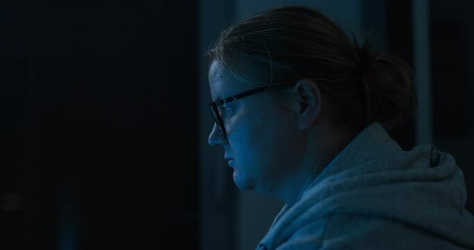 Woman sits in dark room and watches news scrolling Internet. Laptop screen leaves light on adult lady face in empty apartment
