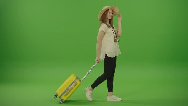 Green Screen. Chroma Key. Pretty Smiling Cheerful Woman Tourist With a Hat on her Head and Yellow Luggage in her Hands Passing by, Winks on Camera. Female on Vacation Trip.