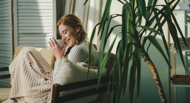 Young relaxed woman in casual outfit reading book and drinking tea while relaxing on sofa in cozy living room at home. Enjoying free time on weekend at home