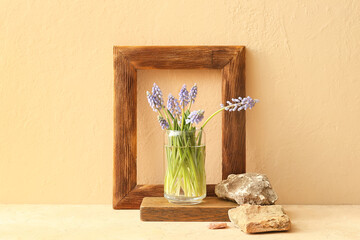 Glass with beautiful Muscari flowers, wooden frame and stones on table near beige wall