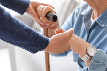 Young caregiver helping senior woman with stick to stand up at home, closeup