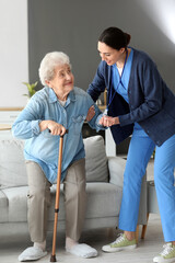 Young caregiver helping senior woman with stick to stand up at home