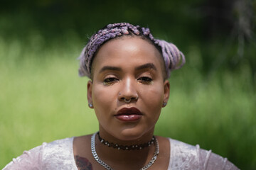 A fresh-faced, dewy person stands in the sunlight, against a defocused backdrop of grass and meadow. Their multiracial, Afro-Latinx complexion is glowing. They are nonbinary, adding more narrative.