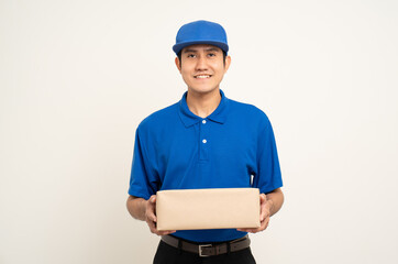 Happy delivery asian man in blue uniform standing holding box parcel cardboard on isolated white background. Smiling male service worker. Delivery courier and shipping service.