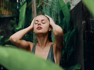 Fototapeta na wymiar A woman takes a shower and washes her head and hair outdoors against the backdrop of tropical plants, palm trees, green banana leaves, summer rain