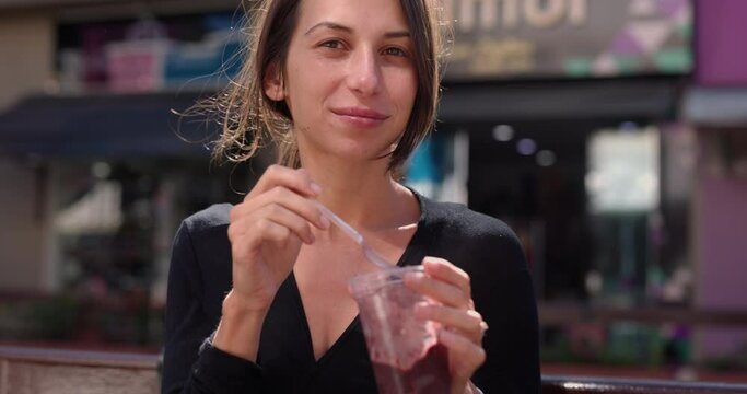 Young woman in 20s eats Acai treat outdoors in summer - looks towards camera