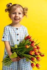 a small blonde on a yellow background with a bouquet of red tulips in her hands, laughing with her eyes closed. the concept of the holiday. vertical photo