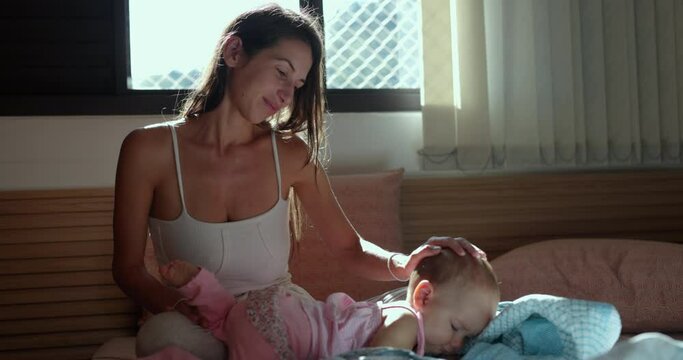 Young mother plays with baby daughter on bed in morning light
