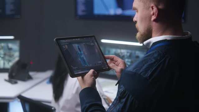 Male officer zooms and controls security cameras, uses tablet in police monitoring center. Security operators working by computers at background. Tracking and monitoring. Concept of safety. Close up.