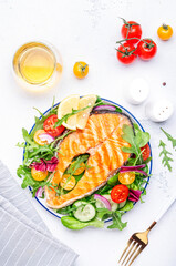 Grilled salmon steak with vegetable salad with tomatoes, arugula, onion  and lettuce on plate, white wine glass, gray table background, top view
