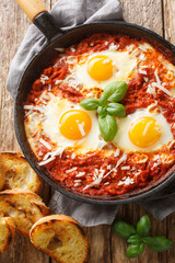 Fried eggs in a spicy tomato sauce with onion and garlic sprinkled with parmesan close-up in a frying pan on a wooden table. Vertical top view from above