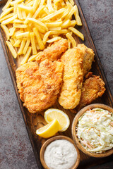 Battered fried fish served with coleslaw, french fries and tartar sauce on a wooden tray on the table. Vertical top view from above