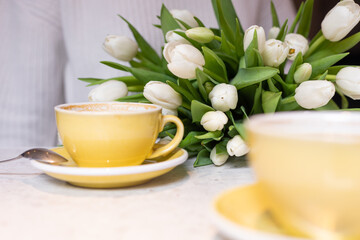 Photography of unrecognized woman with flowers in white dress sitting drinking cup of hot coffee inside