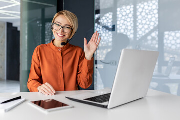 Portrait of successful tech support worker, woman with blonde hair smiling and looking at camera, businesswoman with headset for video call, greeting gesture, inside office with laptop. - obrazy, fototapety, plakaty