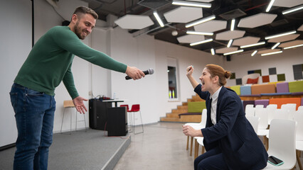A red-haired Caucasian business woman sits in the front row in an empty conference room. Bearded man asks a question in the hall. 