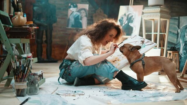 Female artist having fun with her small pet dog sitting on the floor in an art studio