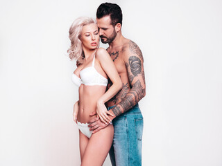 Sexy beautiful woman and her handsome boyfriend. Young passionate couple hugging. Man with nude tattoo torso. Blond model in lace lingerie wearing pure underwear. Posing in studio, near white wall