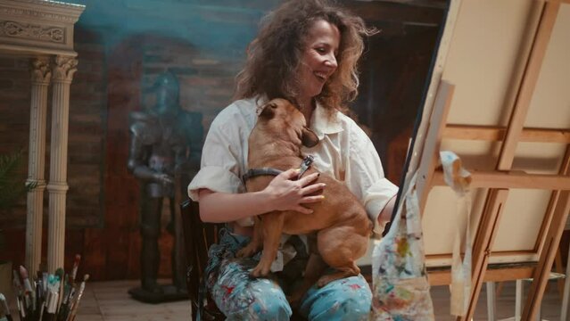Happy female artist is holding her dog in her lap who is kissing her while she is painting in the studio.