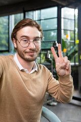 Man in office wearing shirt and glasses smiling and looking at smartphone camera, businessman taking selfie and talking on video call using smartphone, programmer waving hand greeting gesture