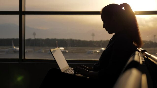 Busy woman works even while waiting for flight at airport lounge. Silhouette of passenger typing at laptop, sitting against window. Bright sunset light shine outside, blurred background, planes parked