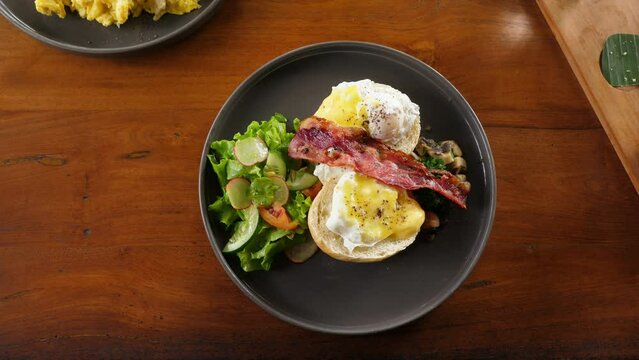 Breakfast is ready, woman put plate on table, split buns with poached eggs served with simple salad, pan-fried slice of bacon and mushrooms. Popular and tasty meal for morning time