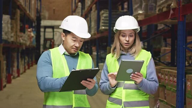 Caucasian woman and asian man with tablet and clipboard, counting and checking goods on warehouse shelves. Focused colleagues wearing safety helmets putting info online to company app.