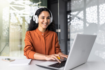 Young successful hispanic woman working in office with headphones, female programmer coding software on laptop smiling, listening to online audio books and podcasts, businesswoman inside office.