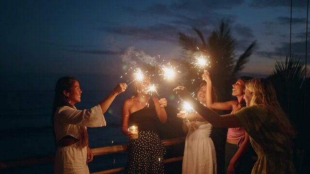 Diverse women dance, laugh with sparklers at seaside tropical hen party at sunset. Young smiling multiracial females have fun at 4th July Independence Day celebration by the sea with sparkling lights