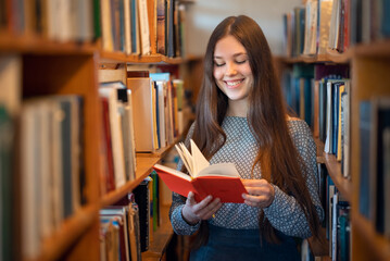 Cheerful female student standing between bookshelves in library of university during break between...