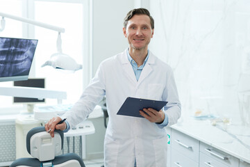Waist up portrait of smiling male dentist looking at camera in dental clinic and holding clipboard, copy space