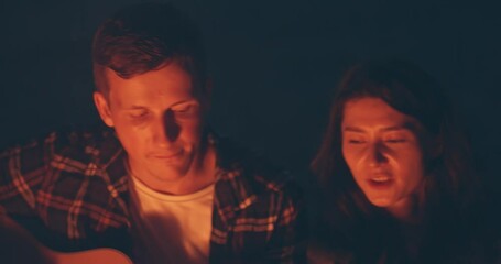 Pretty young couple playing guitar and singing near the bonfire at light night. Social distancing together. two lovers spending time at nature far away from croudy place. Safety way to spend free time - Powered by Adobe
