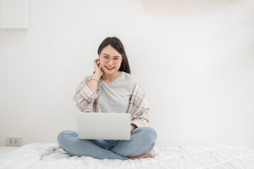 Asian woman looks at the camera using the laptop sitting on the bed in the room