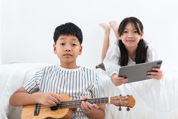 Brother holding an ukulele, sister holding a tablet looking at the camera in the bedroom