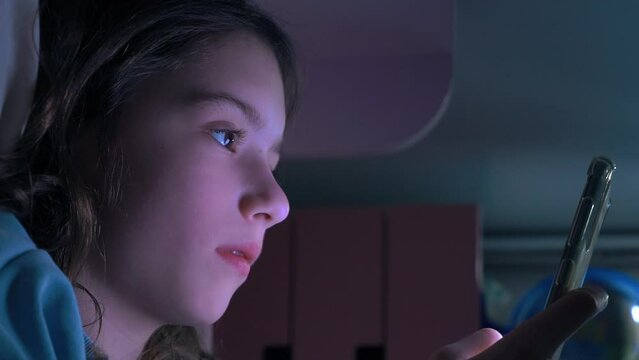 Teen girl scrolling social media texting on smartphone in bed in bedroom at night. Technology, internet, communication and people concept. Dependence on social networks in children. Not healthy sleep.