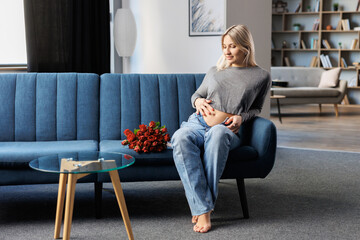 Obraz premium Beautiful pregnant woman sitting on couch in living room near beautiful bouquet