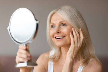 Portrait of beautiful senior woman looking at mirror and smiling, enjoying smooth glow moisturised...