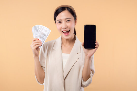 portrait of happy successful confident young asian business woman wearing white jacket holding cash money dollars and credit card standing over beige background. millionaire business, shopping concept