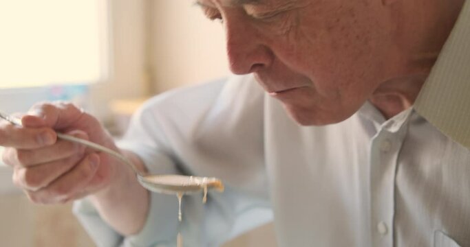 Elderly man eats soup with bread at home in kitchen. Close-up of the face, side view.