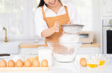 Obraz na płótnie Canvas Young woman standing in kitchen sifting flour for homemade bakery or bread baking ingredient. Asian female chef holding sieve sifted flour preparing for egg mixing in process making pastry or cake.