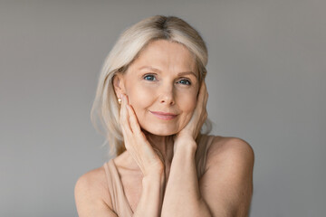 Aged skincare concept. Beautiful senior lady touching her face and smiling, posing on grey wall...