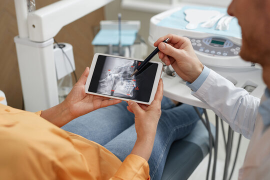 Closeup of male dentist pointing at digital tablet with tooth X-ray image during consultation on surgery in dental clinic