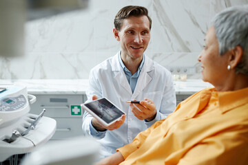 Portrait of smiling male dentist holding digital tablet with teeth x-ray image while explaining dental surgery to patient