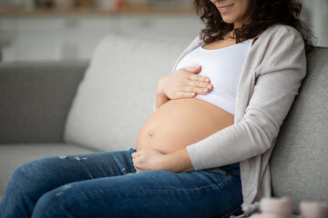 Happy smiling young pregnant woman sitting on couch and embracing belly