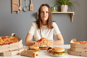 Sad unhappy woman wearing white t- shirt siting at table with fast food in kitchen looking at camera with displeased face felling overeating unhealthy nutrition.