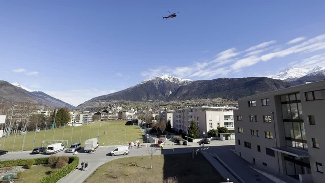 A helicopter transports heavy material through the air in a housing estate. Flight assistants help with loading and unloading
